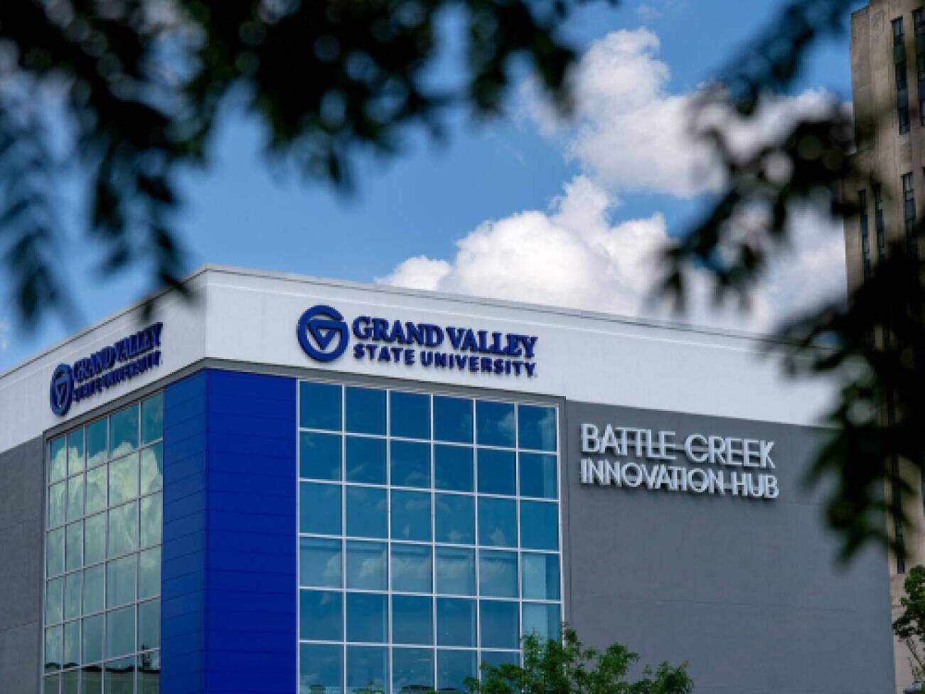 The Grand Valley State University Battle Creek Innovation Hub building is framed by tree branches against a blue sky with clouds.